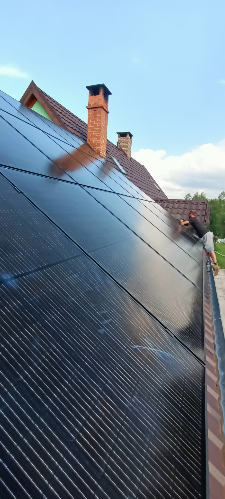 Montaż paneli fotowoltaicznych na dachu domu jednorodzinnego z ceglanym kominem, instalator w okularach ochronnych na drabinie, pochylony nad panelami.