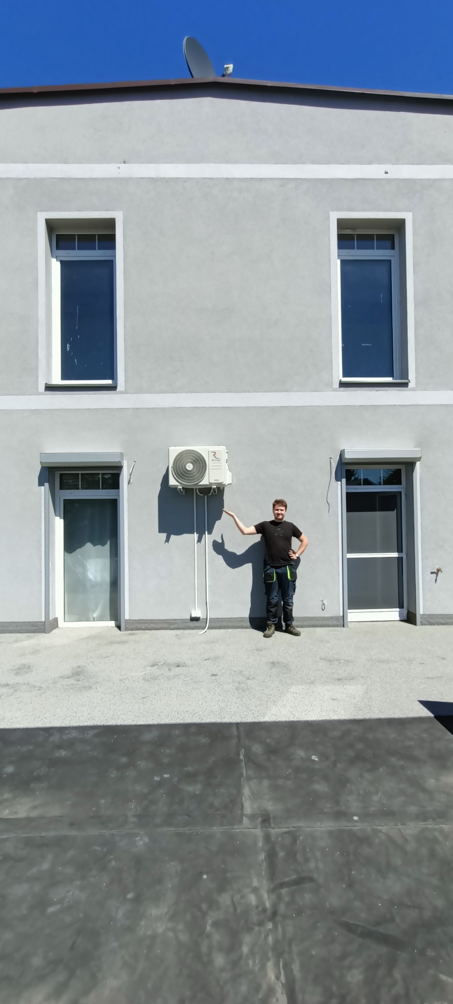 Mężczyzna w ubraniu roboczym prezentuje zamontowaną jednostkę zewnętrzną klimatyzacji na szarej ścianie budynku z dwoma oknami na piętrze i dwoma drzwiami na parterze.