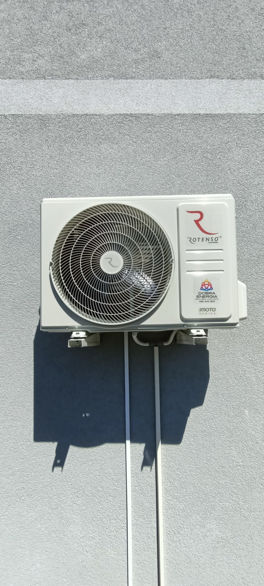 Zewnętrzna jednostka klimatyzacji Rotenso zamontowana na szarej, teksturowanej ścianie budynku, z widocznymi białymi rurami odprowadzającymi skropliny i kablami, oświetlona naturalnym światłem...