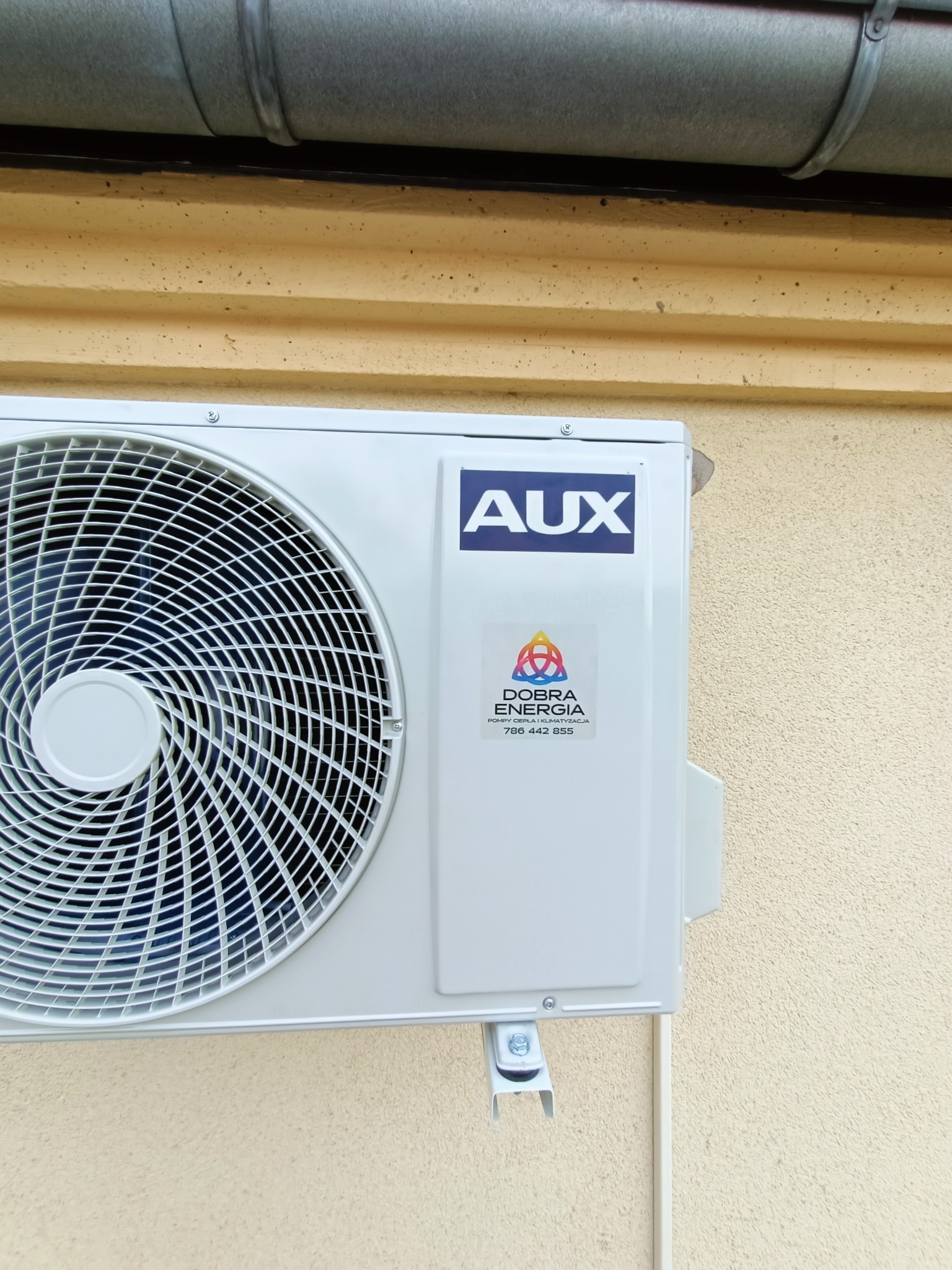 Zewnętrzna jednostka klimatyzacji marki AUX zamontowana na elewacji budynku z widoczną naklejką firmy instalacyjnej 'Dobra Energia' z numerem telefonu, umieszczona pod rynną.