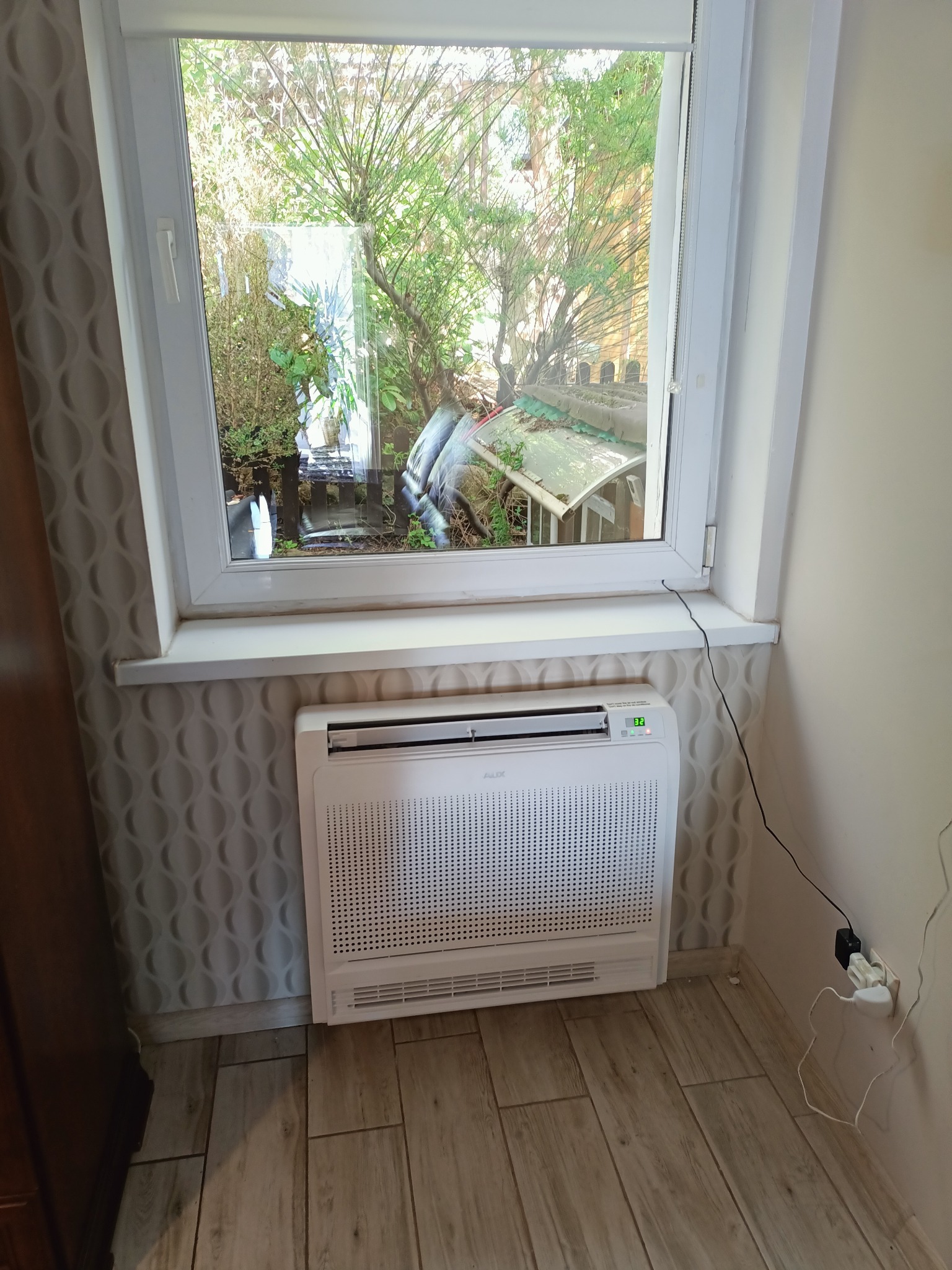 Klimatyzator przypodłogowy w jasnym pomieszczeniu, widok przez okno na zieleń ogrodu, wzorzysta tapeta za urządzeniem.