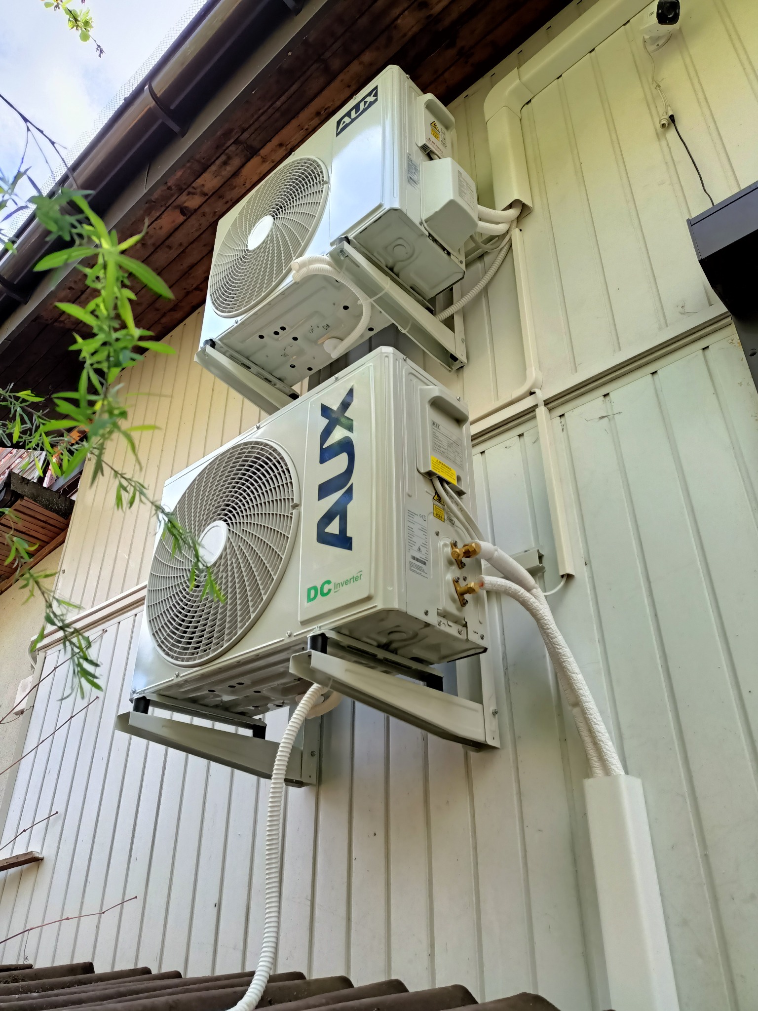 Dwie jednostki zewnętrzne klimatyzacji AUX DC Inverter zamontowane jedna nad drugą na białej ścianie budynku w Leśnej, widoczne przewody instalacyjne i elementy mocujące.