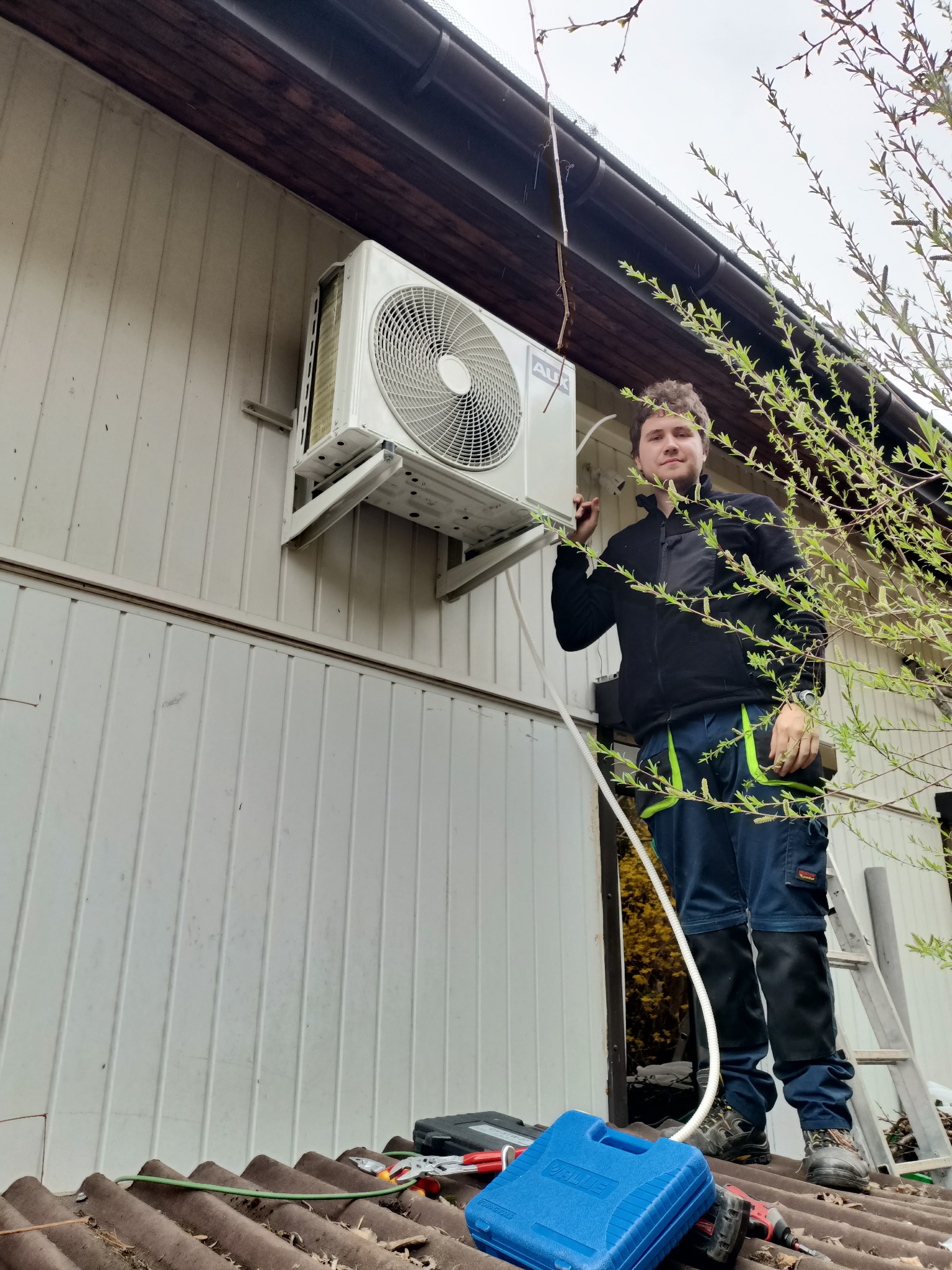 Instalator w czarnej kurtce i roboczych spodniach, stojący na dachu z narzędziami, podłącza białą jednostkę zewnętrzną klimatyzacji do ściany budynku.