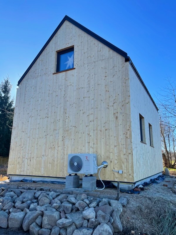 Nowoczesny dom z drewnianą elewacją i zainstalowaną jednostką zewnętrzną klimatyzacji na betonowych bloczkach, widoczne kamienie w ogrodzie i okno z gwiazdą dekoracyjną.