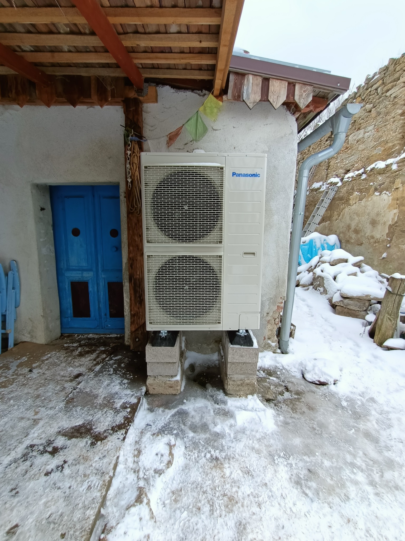 Biała jednostka zewnętrzna Panasonic na betonowych bloczkach, zamontowana na ścianie budynku obok niebieskich drzwi, zimowy krajobraz w tle.