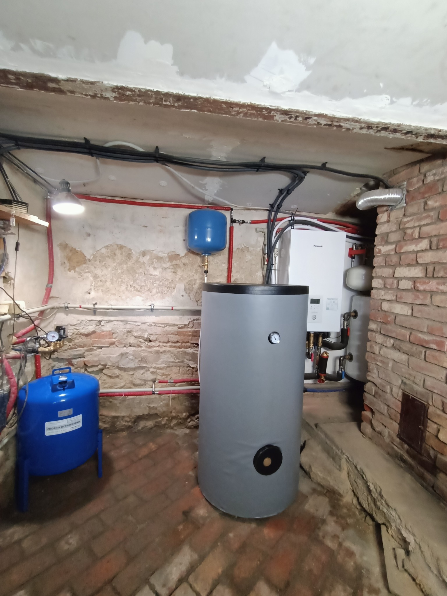 Instalacja pompy ciepła w piwnicy: szary zasobnik ciepłej wody użytkowej, niebieski zbiornik hydroforowy, biała jednostka wewnętrzna pompy ciepła Panasonic na tle ceglanej ściany i zniszczonego...