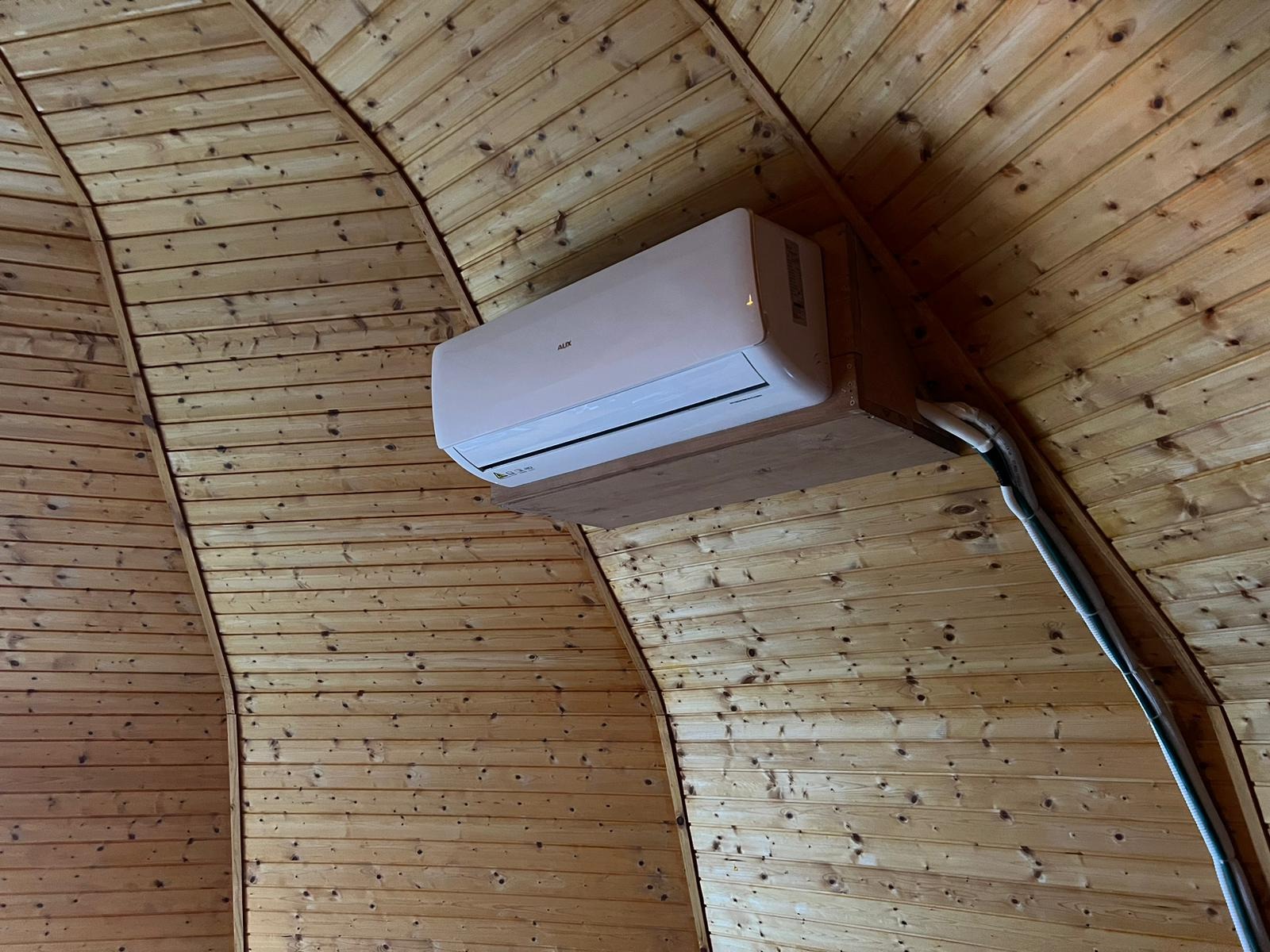 Biały klimatyzator AUX zamontowany na drewnianej ścianie i suficie z boazerii, widoczne rury instalacyjne.