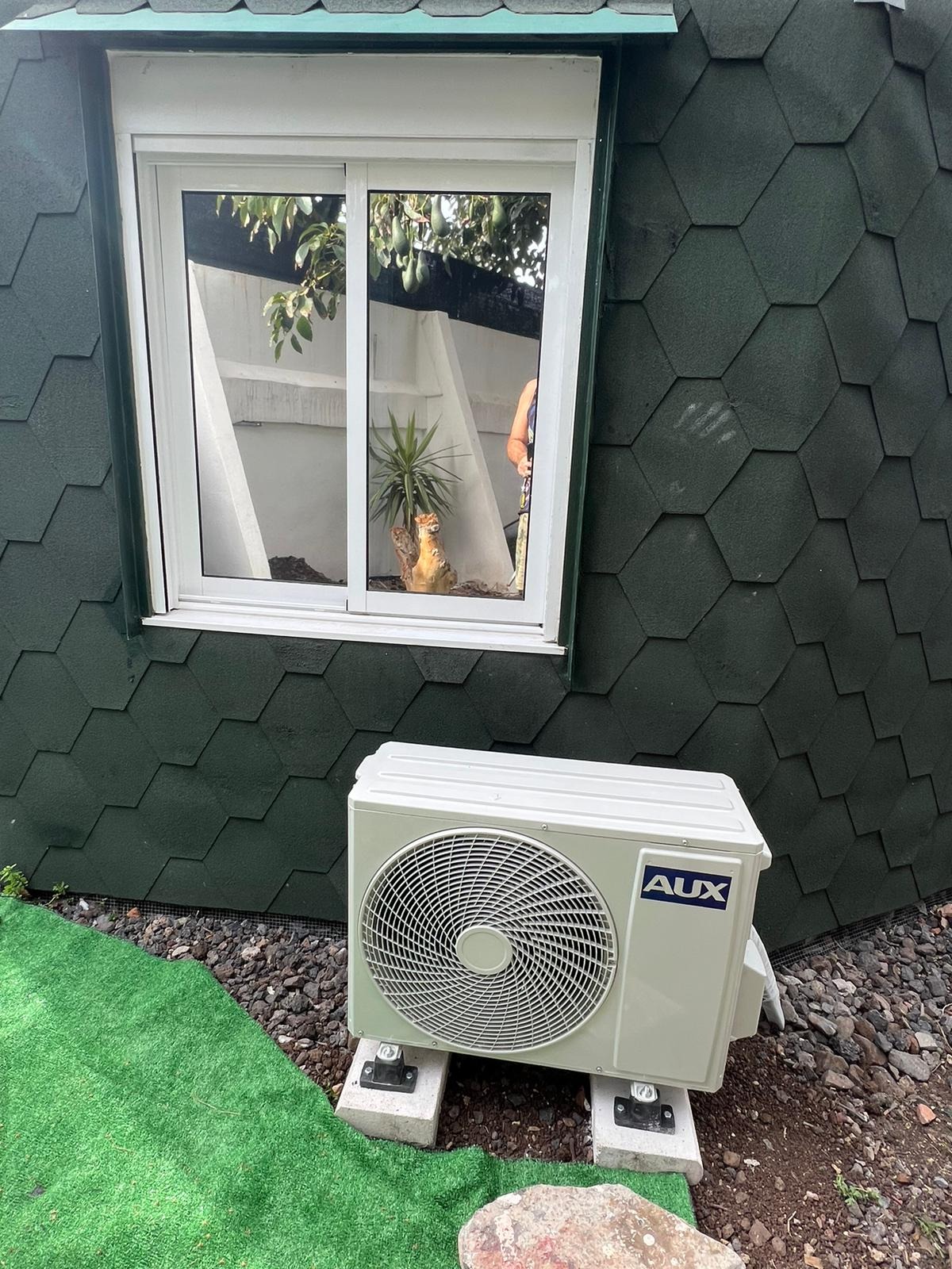 Zewnętrzna jednostka klimatyzacji AUX zamontowana na betonowych bloczkach przed domem z zieloną elewacją imitującą gont, obok sztuczna trawa i kamienie.