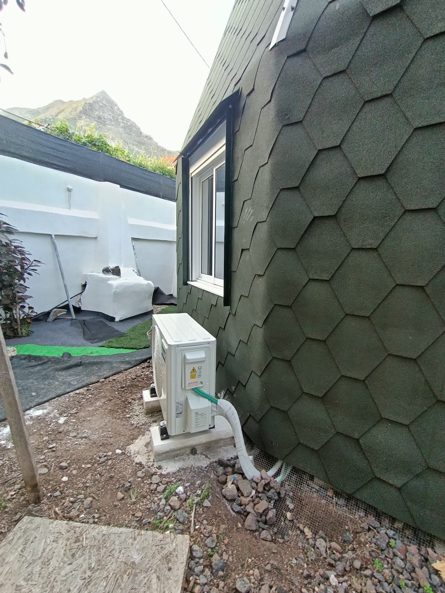 Zewnętrzna jednostka klimatyzacji zamontowana przy budynku pokrytym gontem bitumicznym w kształcie plastra miodu, widok na górę w tle.