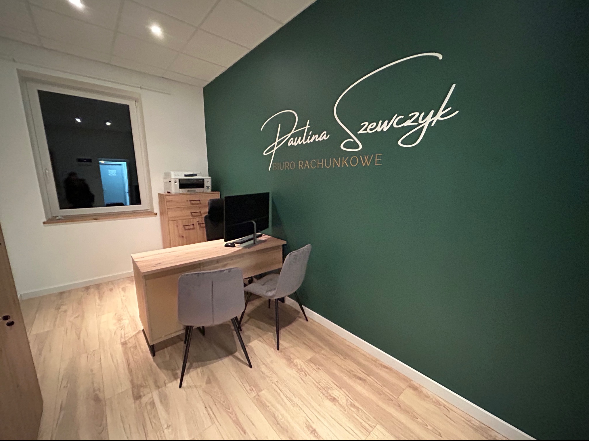 Wnętrze biura z zieloną ścianą, na której widnieje białe, ozdobne logo firmy 'Paulina Szewczyk Biuro Rachunkowe', biurko z komputerem i dwoma krzesłami.