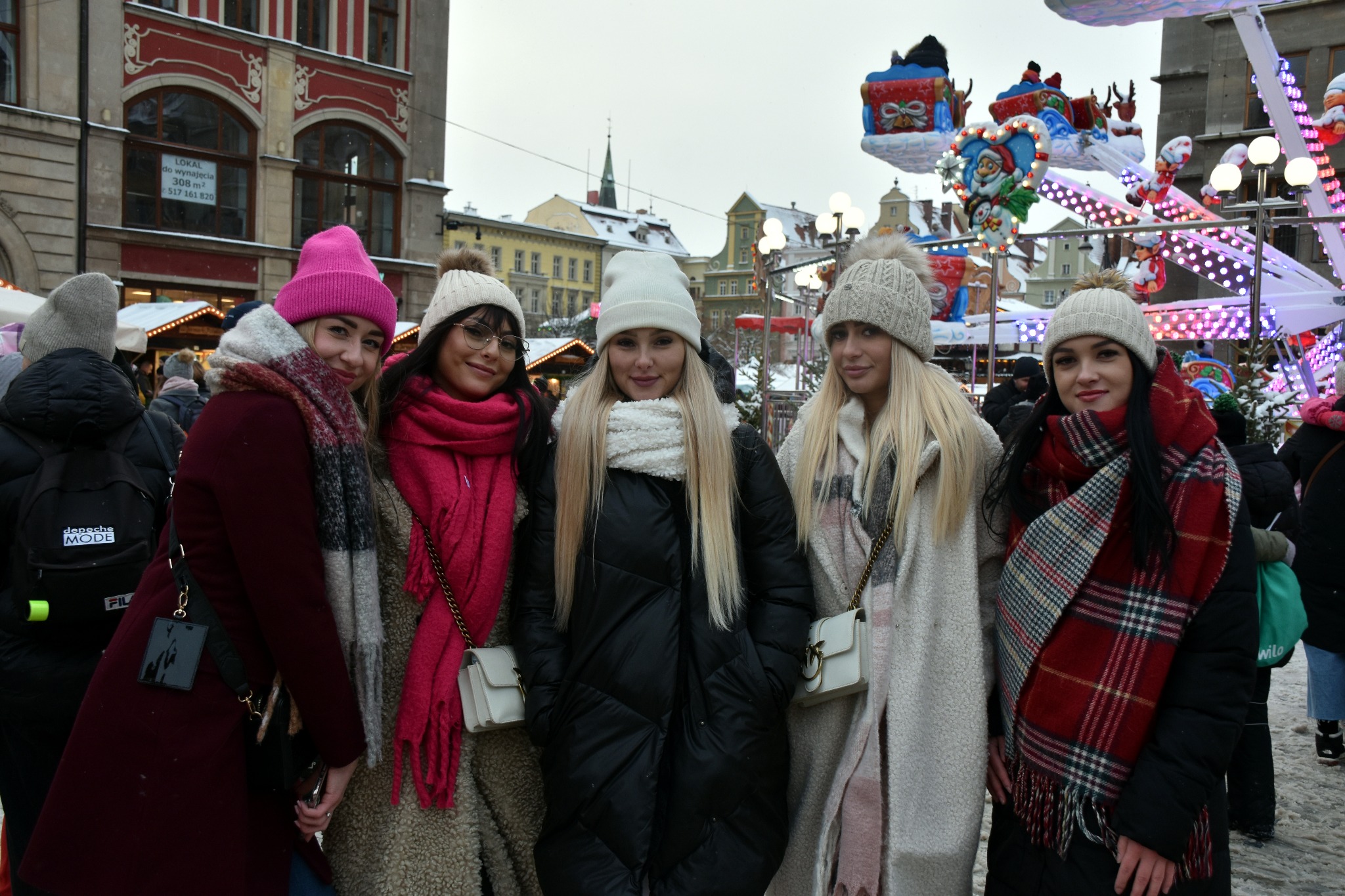 Grupa pięciu młodych kobiet w zimowych ubraniach pozuje na tle świątecznego jarmarku z karuzelą i oświetlonymi budynkami w tle.