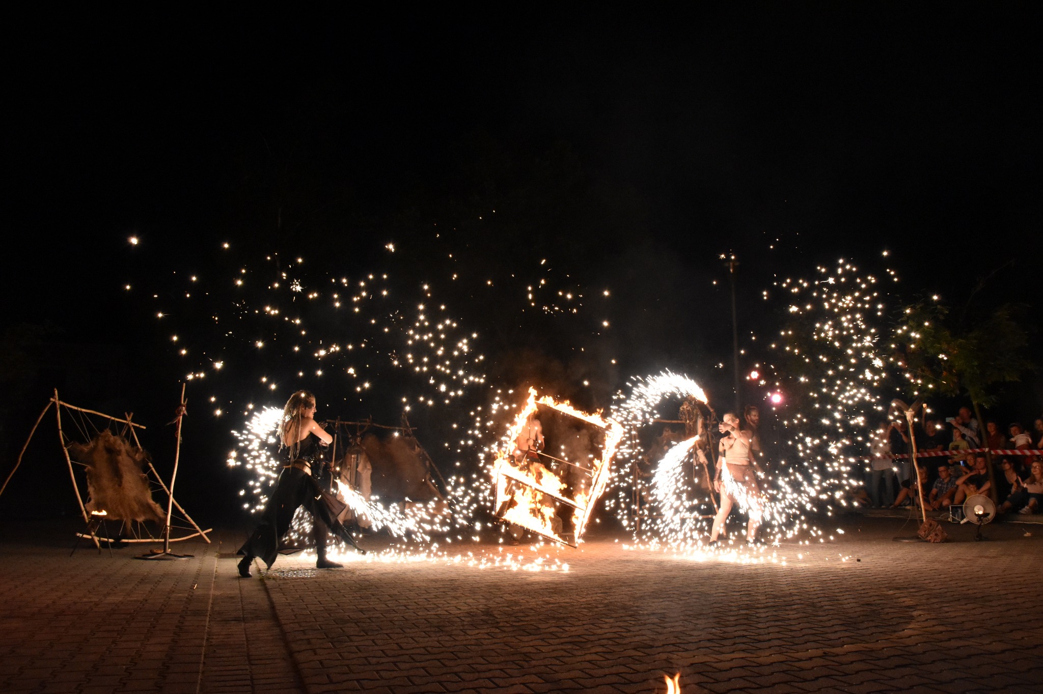 Pokaz fire show w nocy: dwie kobiety w kostiumach tańczą z płonącymi rekwizytami, iskry i ogień oświetlają publiczność w tle.