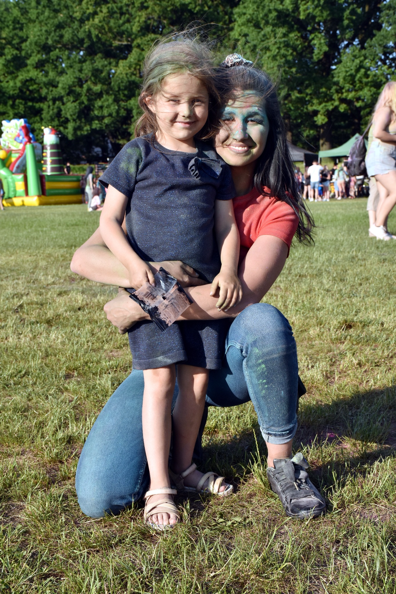 Uśmiechnięta kobieta obejmuje dziewczynkę z pomalowanymi twarzami na tle parku z dmuchanym zamkiem.