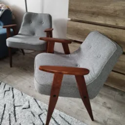 Dwa szare fotele tapicerowane z drewnianymi podłokietnikami i nogami w stylu retro, ustawione na dywanie w salonie z drewnianą komodą w tle.