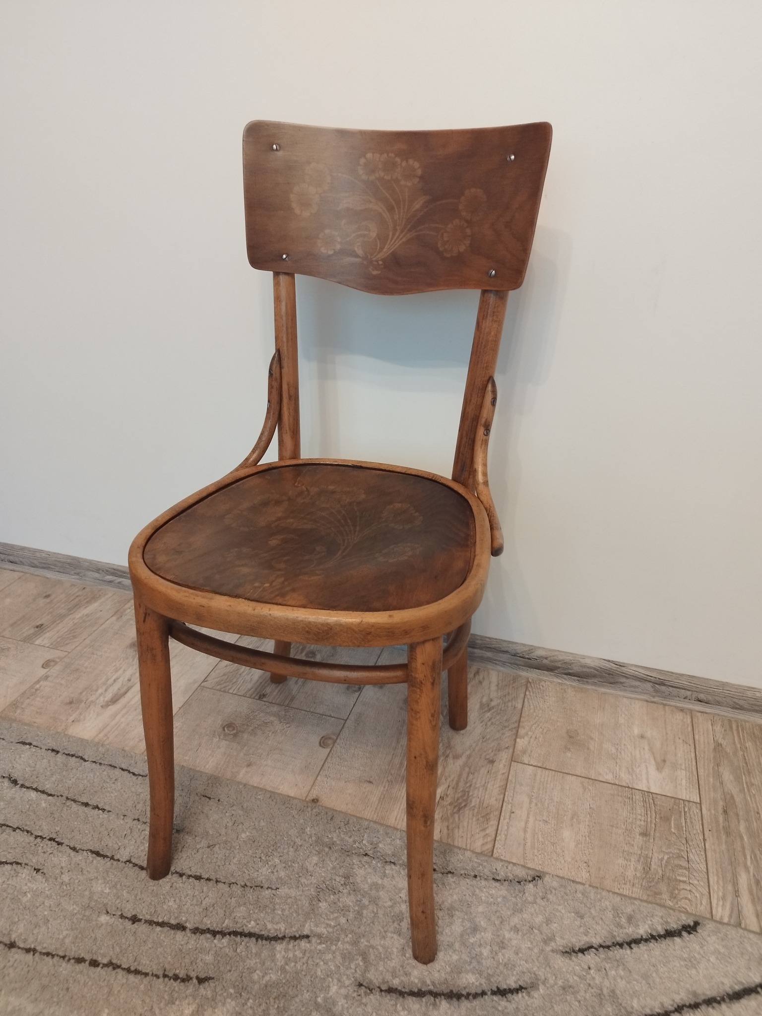 Drewniane krzesło gięte w stylu retro, z delikatnym kwiatowym wzorem na oparciu i siedzisku, stojące na dywanie w pokoju.
