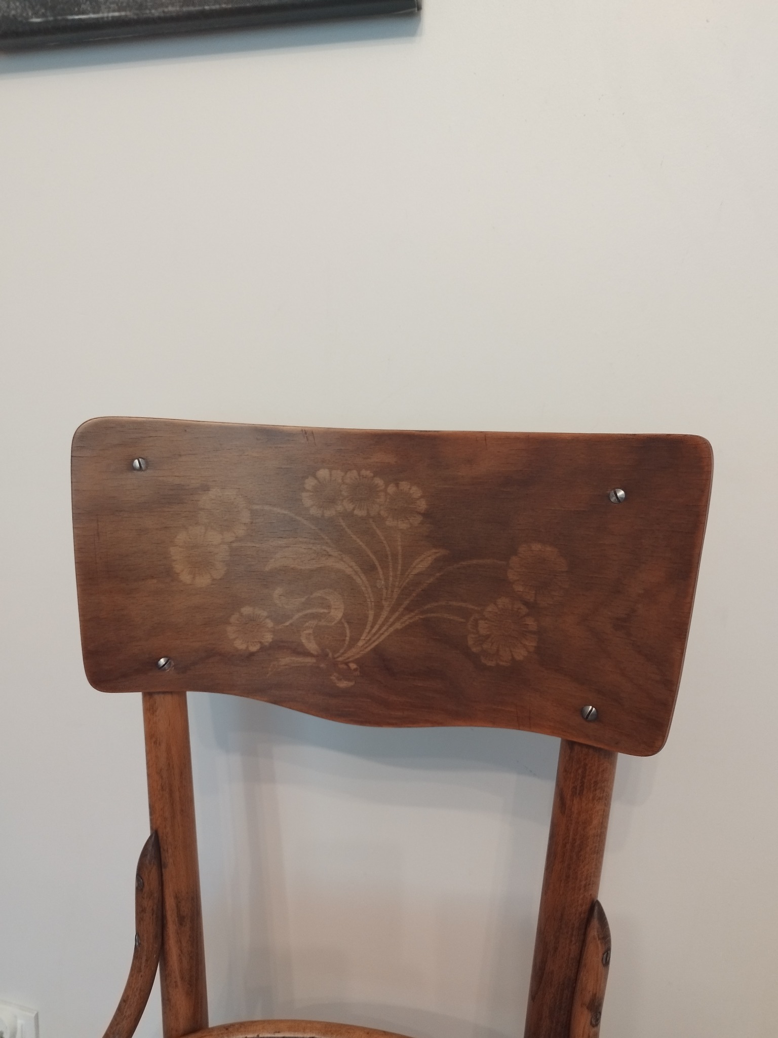 Drewniane krzesło z oparciem zdobionym motywem kwiatowym, mocowane na cztery śruby, na tle białej ściany.