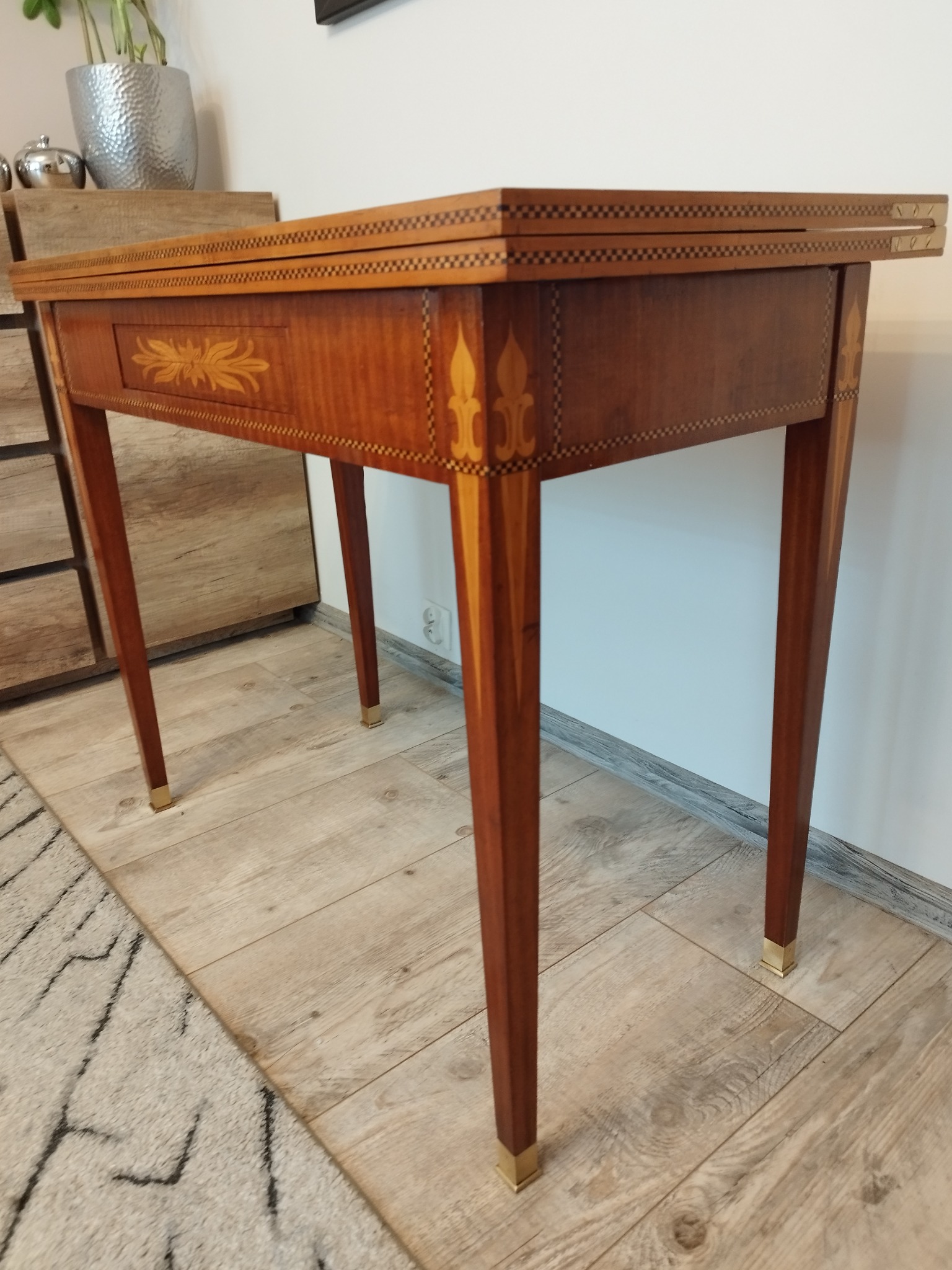 Rozkładany, fornirowany stolik z intarsjami, z dekoracyjnymi motywami kwiatowymi i geometrycznymi na blacie i nogach, wykończony metalowymi stopkami.