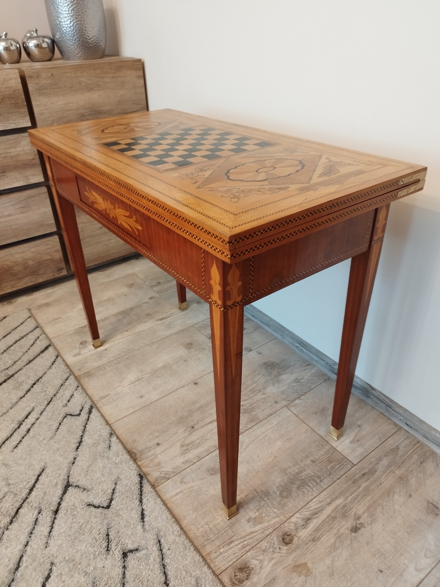 Rozkładany, drewniany stolik do gier z szachownicą i dekoracyjnymi intarsjami, ustawiony na drewnianej podłodze obok wzorzystego dywanu.