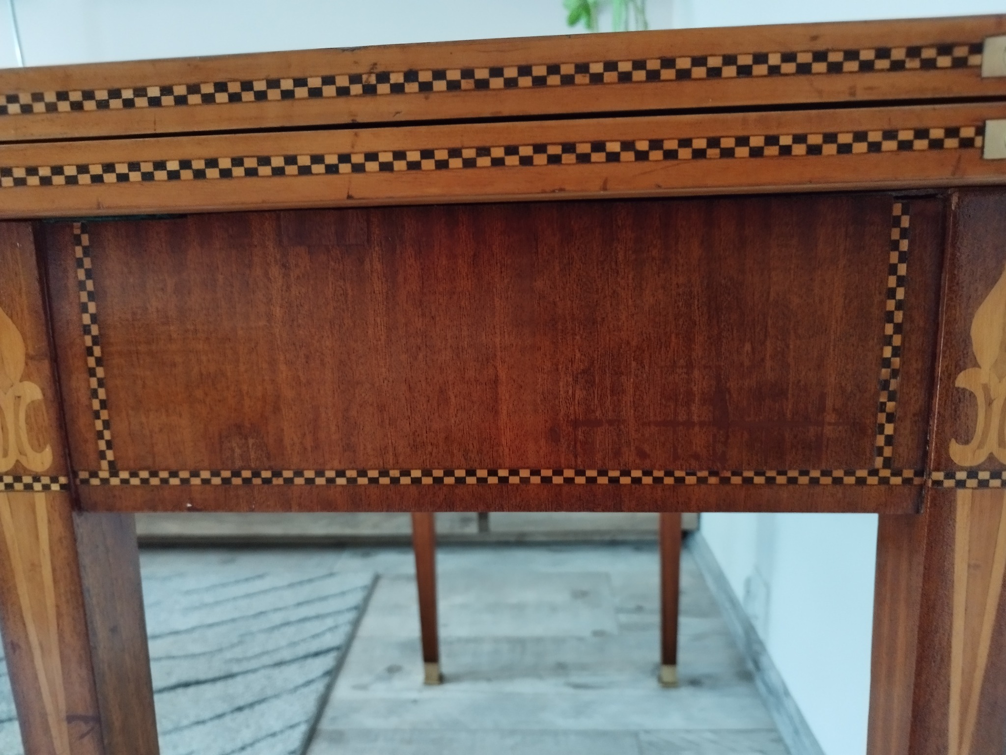 Fragment odrestaurowanego, drewnianego stolika z dekoracyjnymi intarsjami w motywy szachownicy i roślinne, widoczne detale wykończenia i forniru.