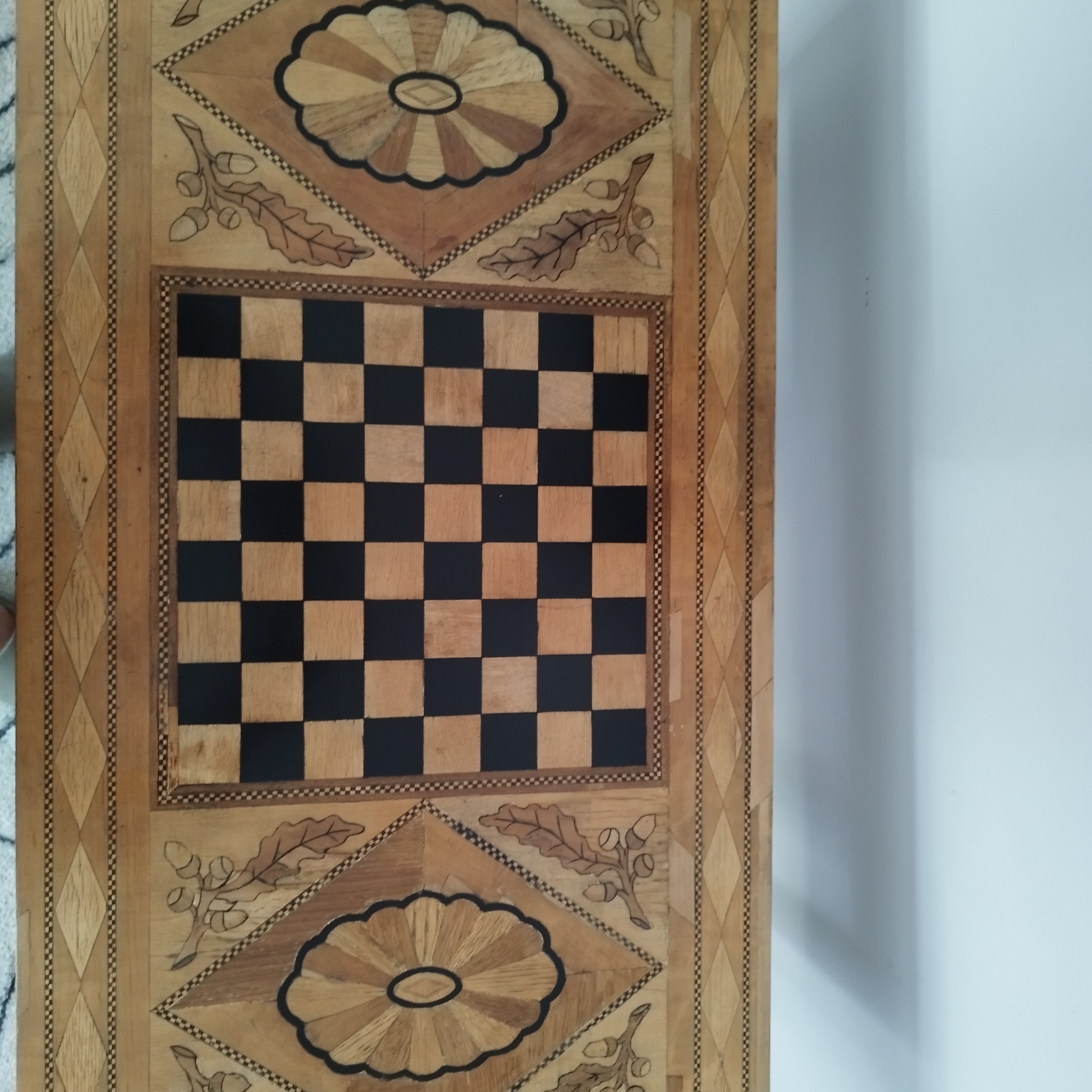 Drewniany blat stołu z intarsją, przedstawiający szachownicę otoczoną motywami kwiatowymi i liśćmi dębu.