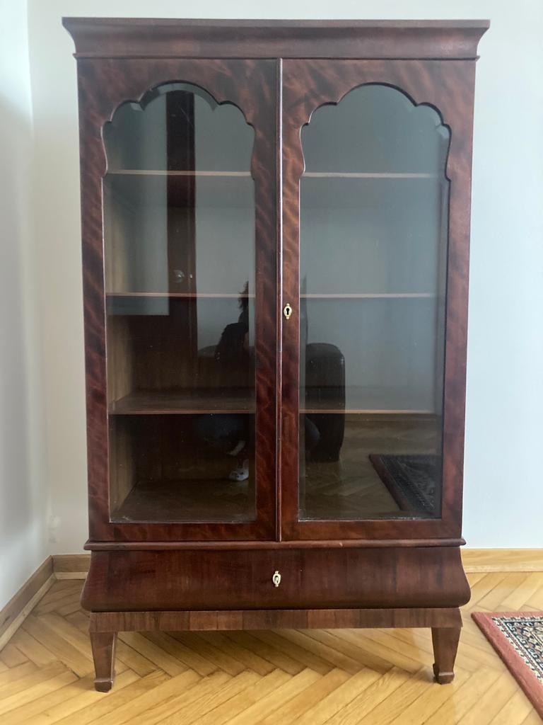 Antyczny, dwudrzwiowy kredens z ciemnego drewna z przeszkleniami w stylu art deco i pojedynczą szufladą, stojący na parkiecie.