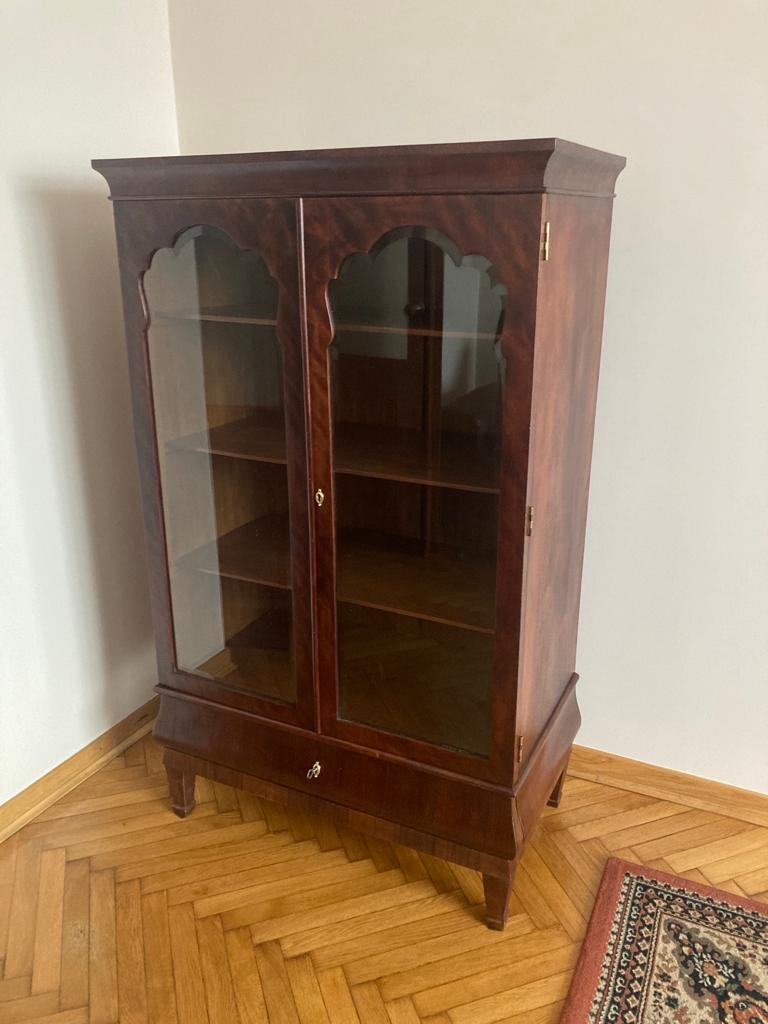 Masywna, ciemnobrązowa witryna z dwoma przeszklonymi drzwiczkami, ozdobnymi wycięciami w górnej części i małym mosiężnym zamkiem, stoi na parkiecie w rogu pokoju obok dywanu.