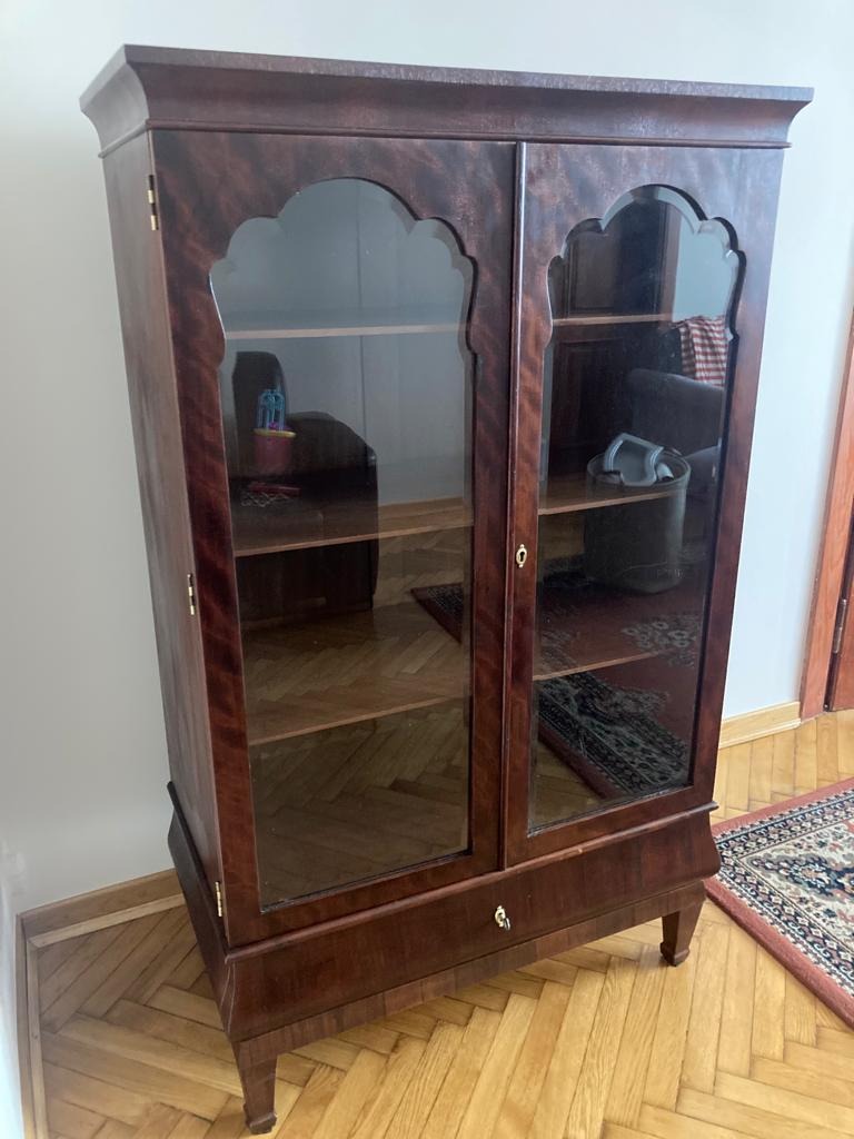 Dwudrzwiowa, fornirowana mahoniem witryna z szybami o łukowatym kształcie, stojąca na parkiecie w jodełkę, z widocznym fragmentem dywanu.