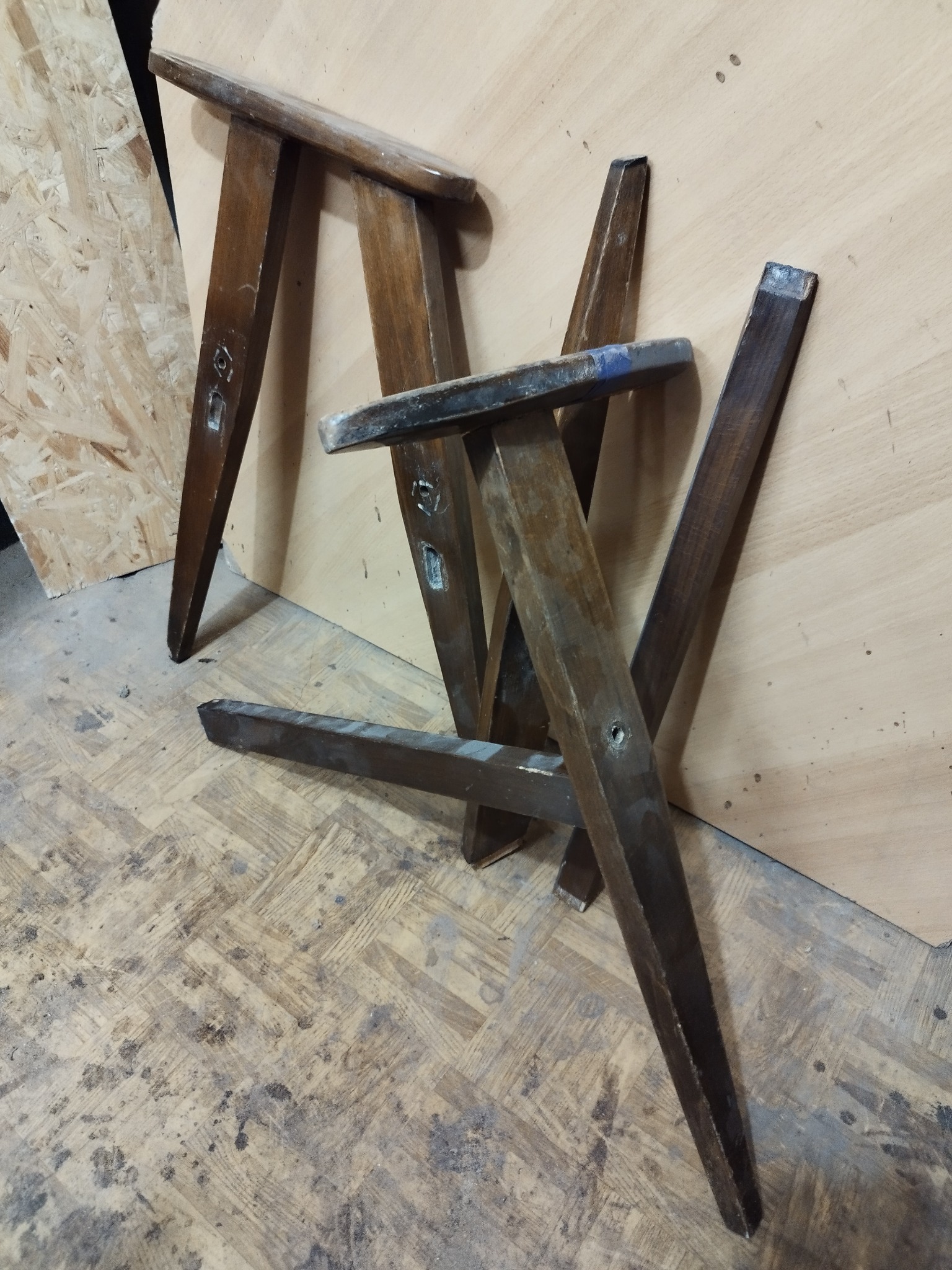 Rozłożone, brązowe, drewniane nogi meblowe z widocznymi śladami użytkowania i otworami na śruby, leżące na podłodze z wzorem parkietu.