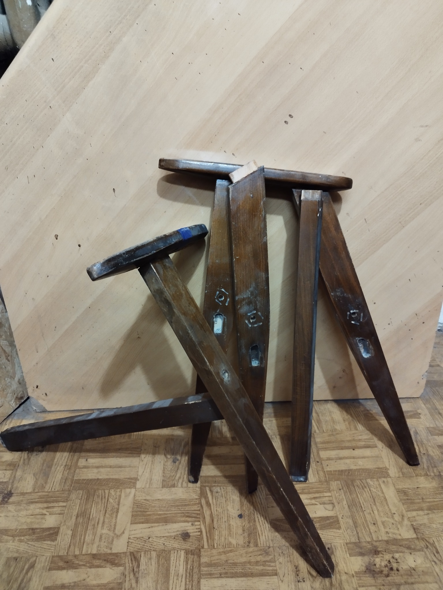 Nierówne nogi od krzeseł lub stołków w ciemnym odcieniu brązu, leżące na podłodze z wzorem parkietu i na tle płyty meblowej.