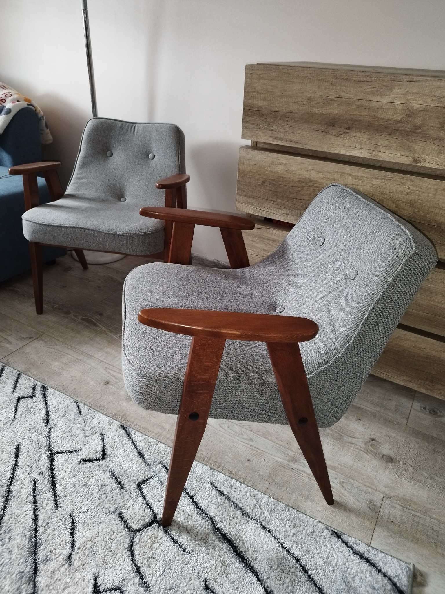 Dwa szare fotele tapicerowane z drewnianymi podłokietnikami i nogami w stylu retro, ustawione na dywanie w salonie z drewnianą komodą w tle.