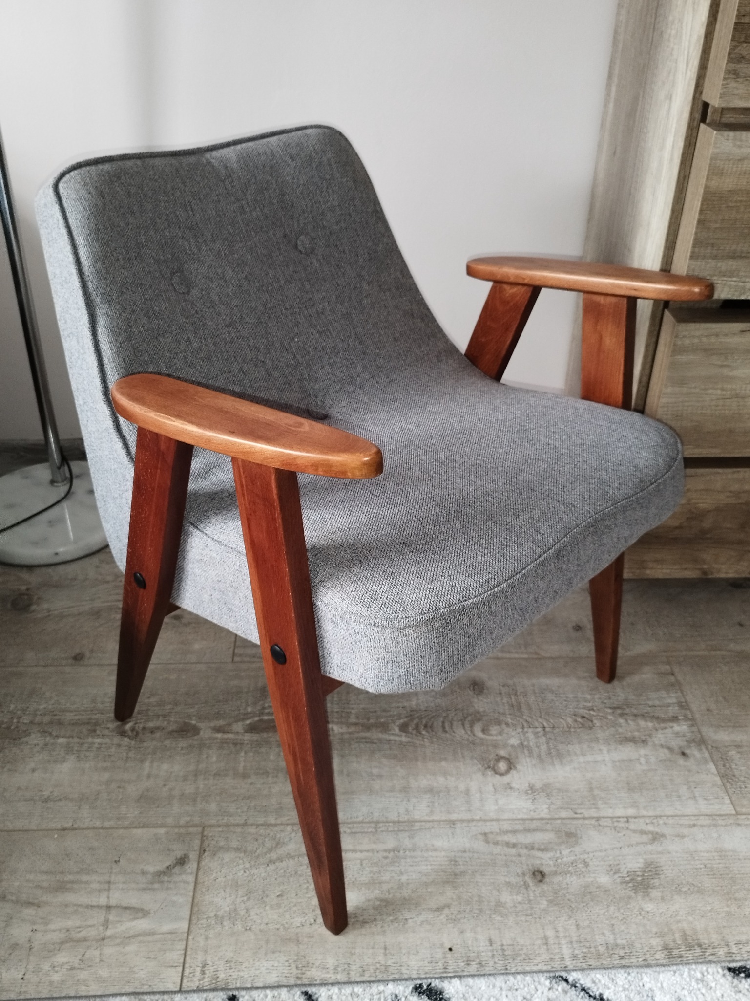 Szary fotel tapicerowany w stylu retro z drewnianymi podłokietnikami, widoczne guziki tapicerskie i drewniane nogi, ustawiony na jasnej podłodze.