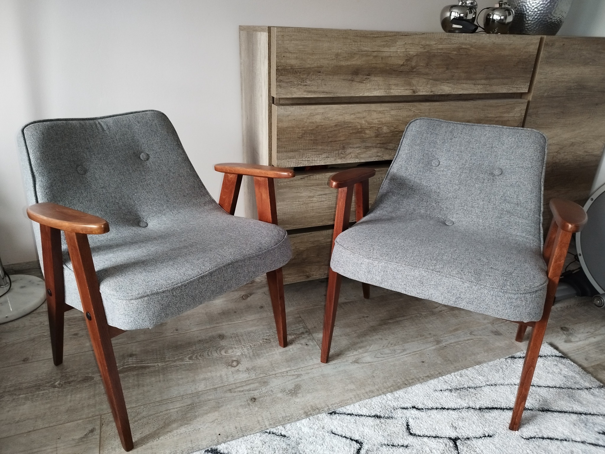 Dwa szare fotele z drewnianymi podłokietnikami i nogami, ustawione na wzorzystym dywanie, z komodą w tle i srebrnymi dekoracjami na niej.