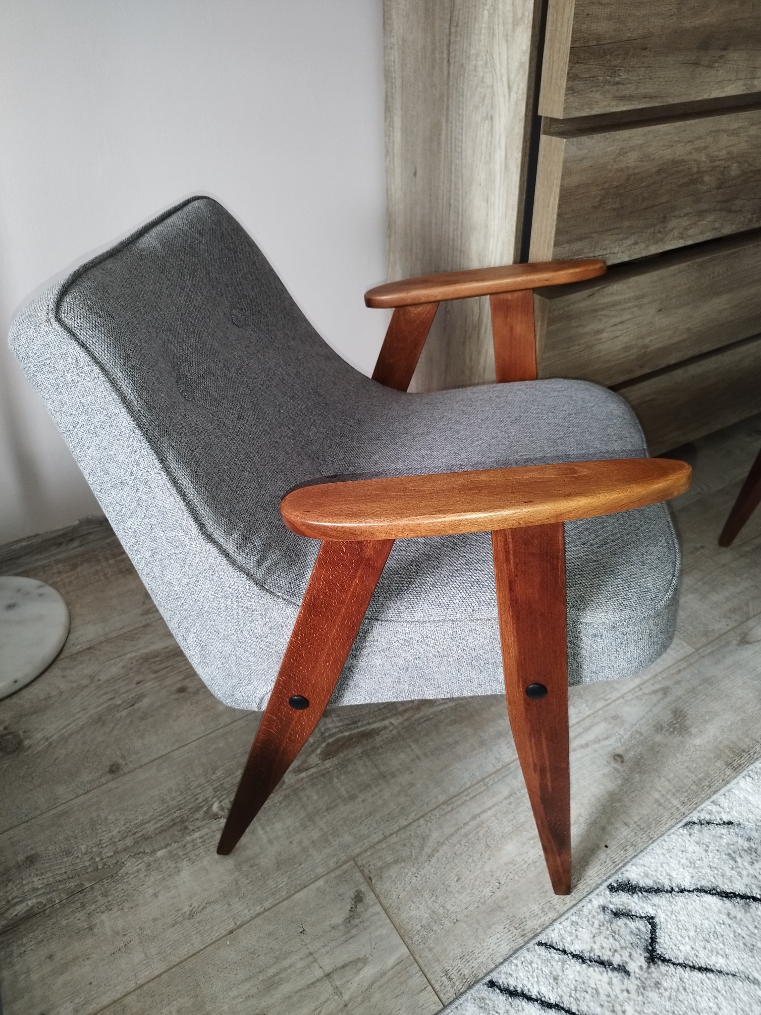 Tapicerowany fotel w stylu retro z drewnianymi podłokietnikami i nogami, na tle drewnianej komody i marmurowego stolika.