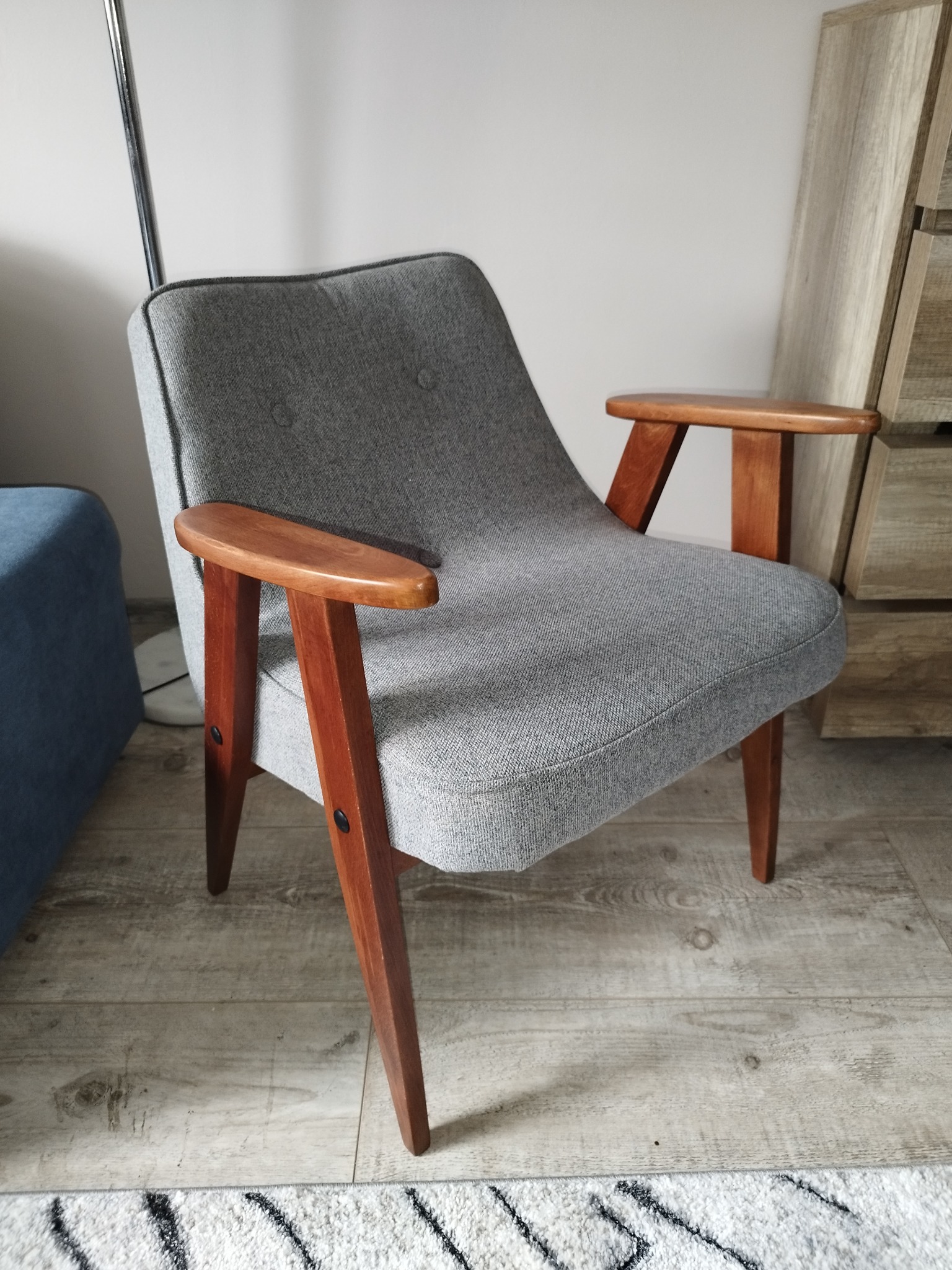 Szary fotel tapicerowany w stylu retro z drewnianymi podłokietnikami i nogami, ustawiony na drewnianej podłodze z fragmentem wzorzystego dywanu w tle.
