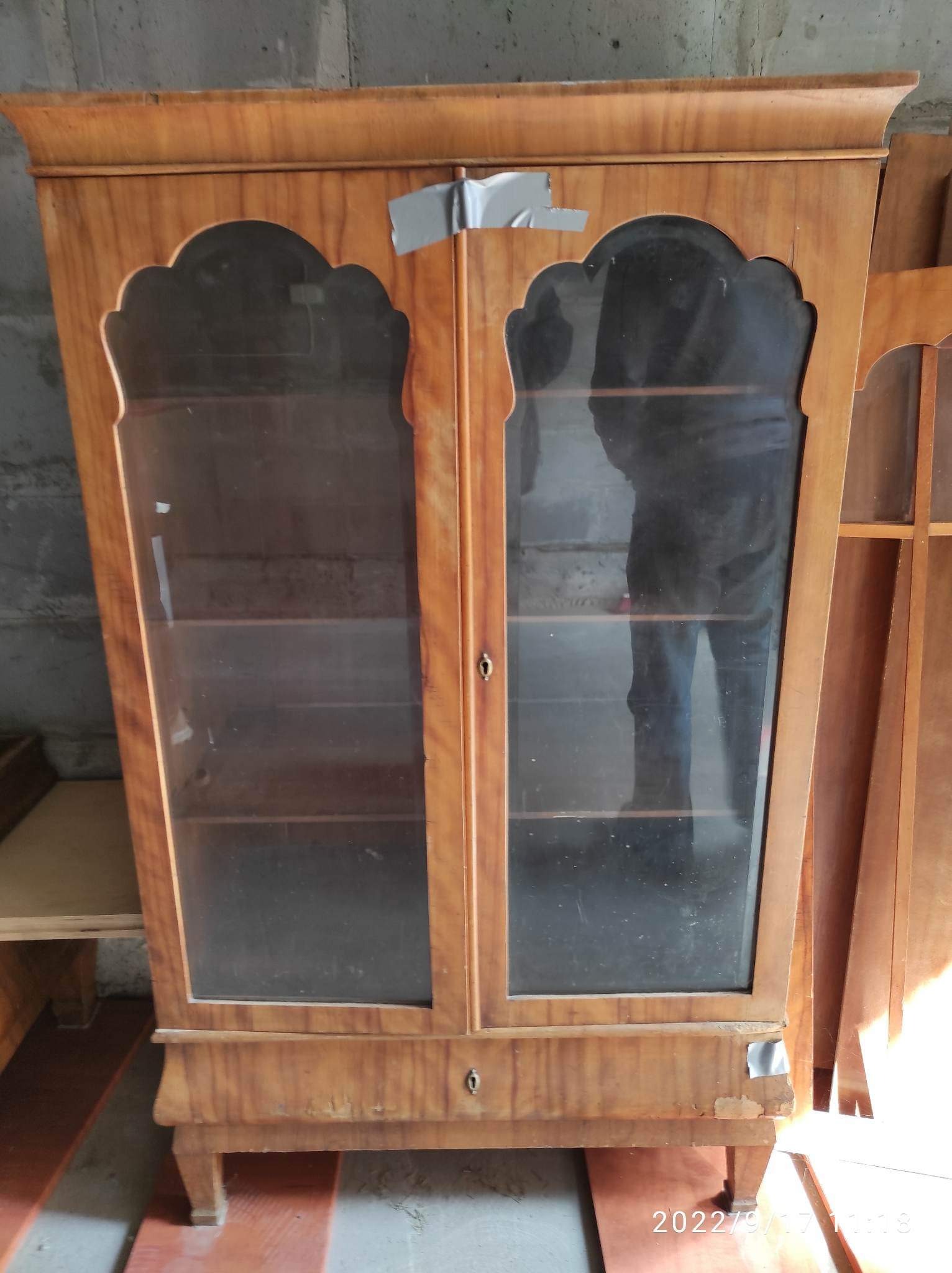 Stara, drewniana witryna z dwoma przeszklonymi drzwiczkami, częściowo zaklejona taśmą, ukazująca ślady użytkowania i potrzebę renowacji.