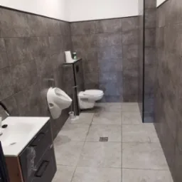 Nowoczesna toaleta publiczna z szarymi płytkami, umywalką z szafką i pisuarami. Minimalistyczny design, jasne oświetlenie, czyste i przestronne wnętrze.