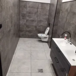 Nowoczesna łazienka po remoncie: szare płytki na ścianach i podłodze, biała armatura, umywalka z szafką. Widoczny odpływ liniowy w podłodze.