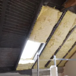 Ocieplenie poddasza wełną mineralną widoczne od wewnątrz, z folią paroizolacyjną i elementami konstrukcji dachu, z oknem dachowym i drabiną w tle.