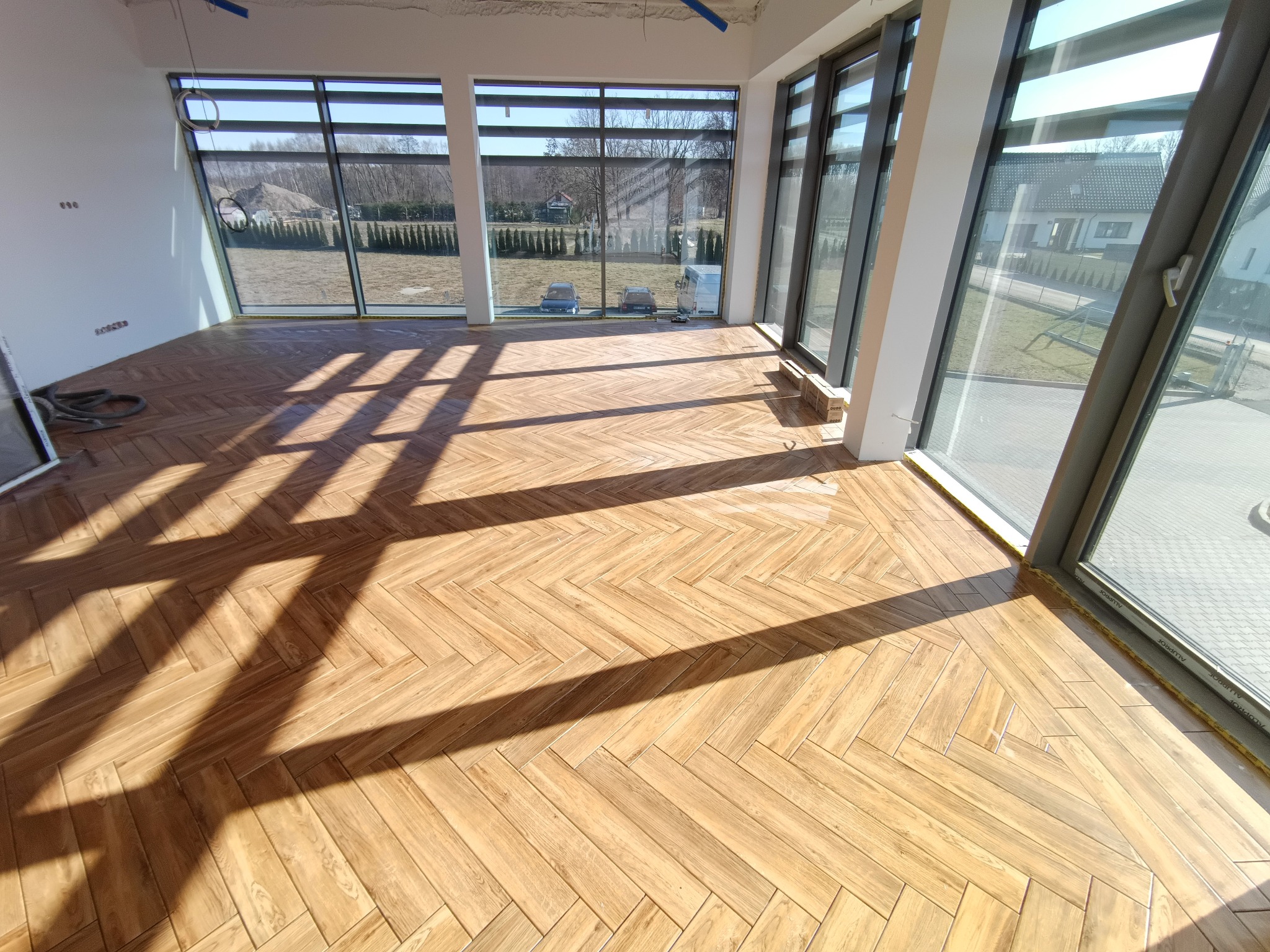 Jasne panele ułożone w jodełkę w przestronnym, jasnym pokoju z dużymi oknami. Widoczne niedokończone prace wykończeniowe przy suficie i ścianach.