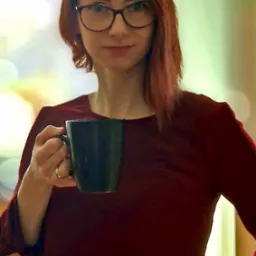 Portret uśmiechniętej kobiety z rudymi włosami w okularach, trzymającej czarny kubek w dłoni z złotym pierścionkiem, na rozmytym tle.