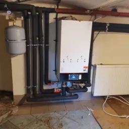 Nowoczesny system grzewczy z białym kotłem i czarnymi rurami izolowanymi, szarym zbiornikiem wyrównawczym i grzejnikiem panelowym na tle beżowej ściany i brudnej podłogi z płytkami.