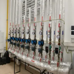 Rząd rur instalacji grzewczej, zaizolowanych folią aluminiową, z licznymi zaworami, manometrami i pompami obiegowymi, obok dw&oacute;ch kotł&oacute;w grzewczych i zbiornika wyr&oacute;wnawczego Reflex w pomieszczeniu...