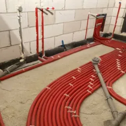 Czerwone rury ogrzewania podłogowego ułożone w pętle na wylewce, widoczne przyłącza do ściany i odpływ podłogowy.