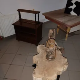 Drewniany stolik kawowy z blatem w kształcie plastra drewna, na którym stoi stara lampa naftowa, w tle komoda i stół z obrazem.