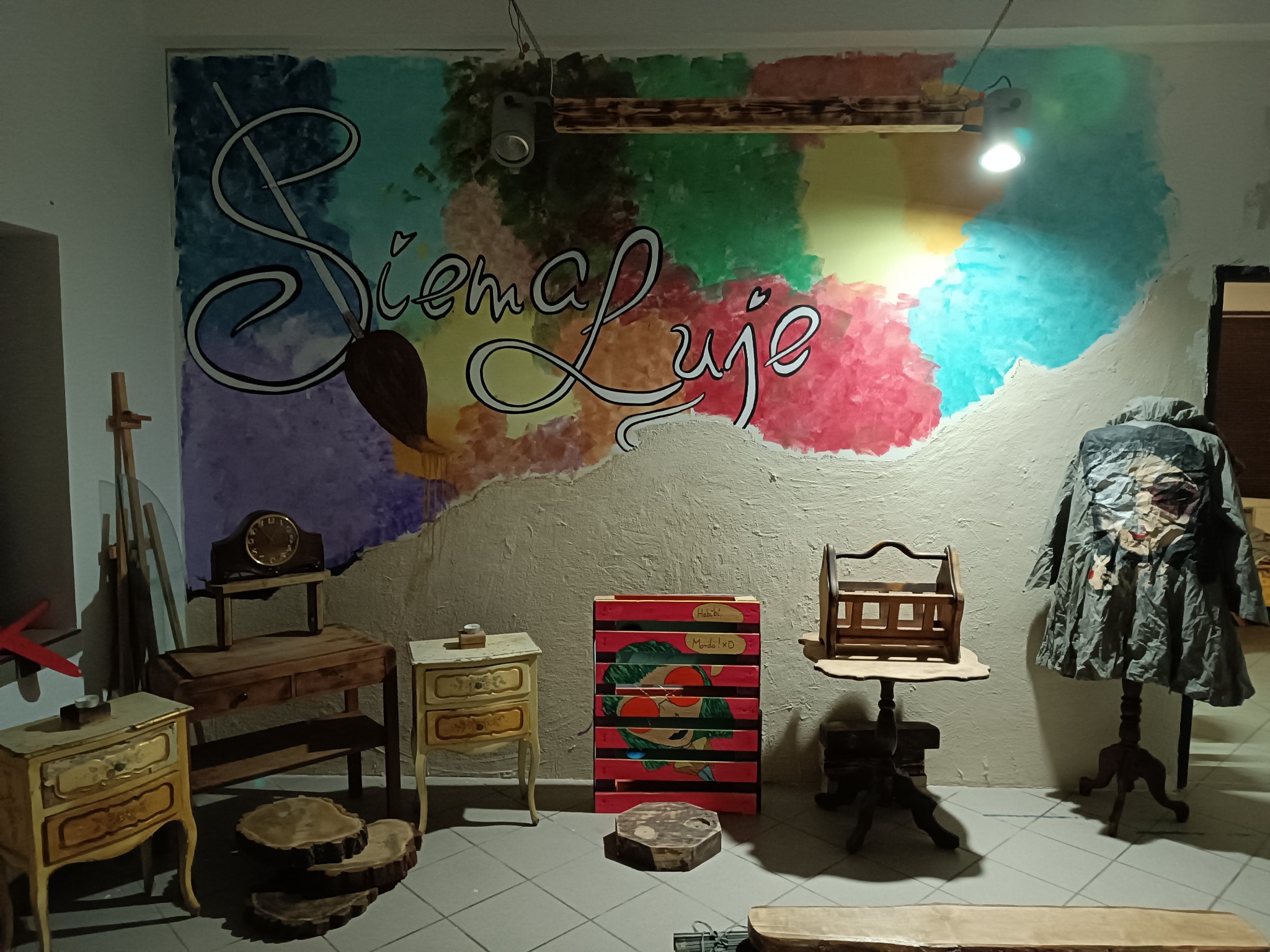 Wnętrze pracowni artystycznej z ręcznie malowaną ścianą z napisem 'Siema Luje' i motywami malarskimi, prezentujące odrestaurowane meble, drewniane elementy dekoracyjne i manekin z artystycznym...