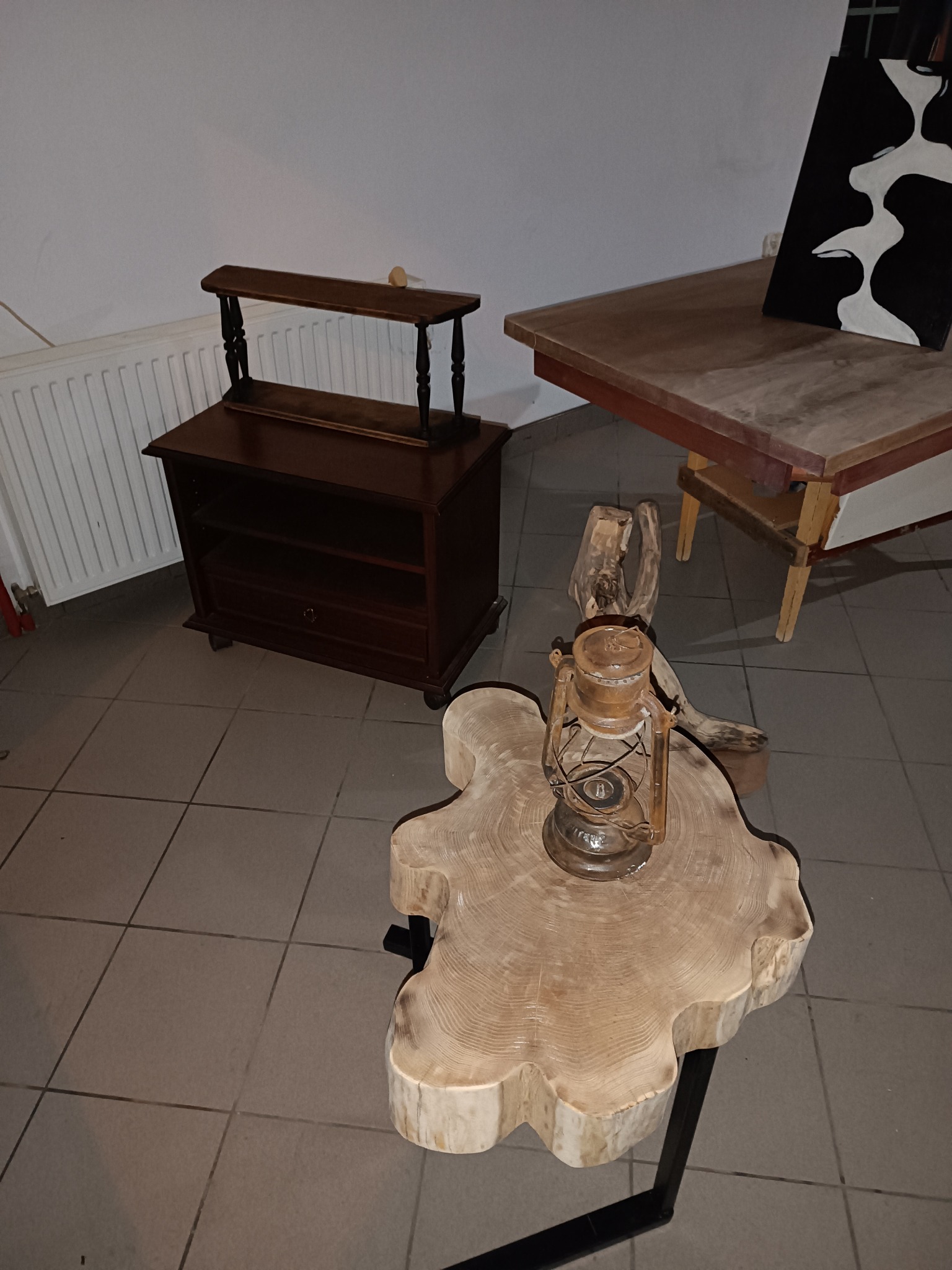 Drewniany stolik kawowy z blatem w kształcie plastra drewna, na którym stoi stara lampa naftowa, w tle komoda i stół z obrazem.