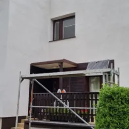 Dwukondygnacyjny dom z białymi ścianami po tynkowaniu, z drewnianymi oknami i fragmentem elewacji wykończonym drewnem. Na balkonie widoczne rusztowanie z przygotowanymi materiałami budowlanymi.