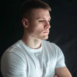 Portret młodego mężczyzny z krótkimi włosami, ubrany w białą koszulkę, z zamyśloną miną, na ciemnym tle.