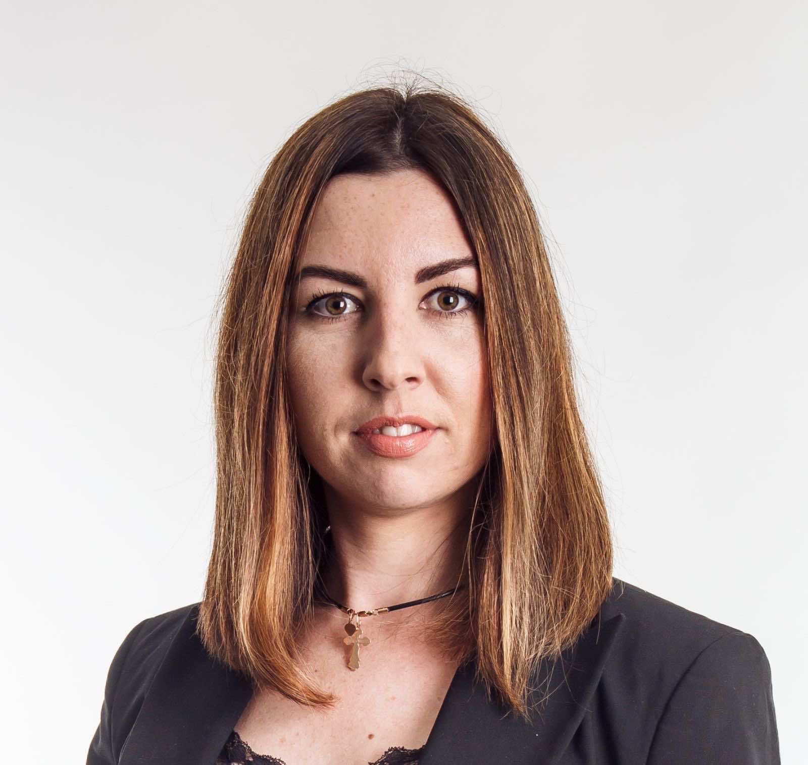 Portret kobiety z brązowymi włosami w czarnej marynarce i z delikatnym naszyjnikiem z krzyżykiem, na jasnym tle.