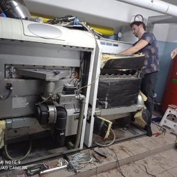 Kupiec - Therm s.c. - Mężczyzna w trakcie prac serwisowych przy otwartym piecu grzewczym, widoczne elementy izolacyjne, narzędzia na podłodze i czerwony zbiornik wyrównawczy.